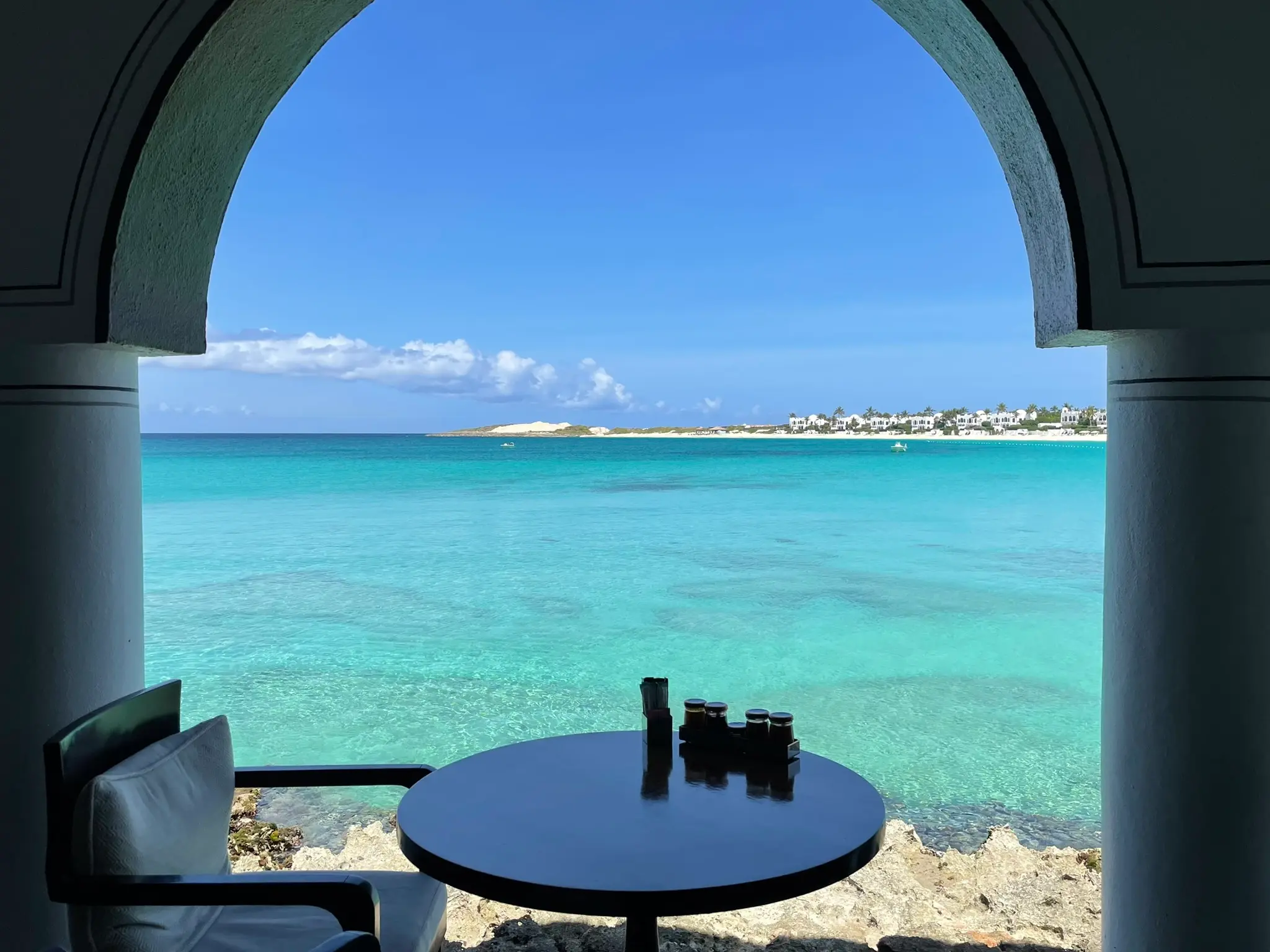 Table and chair by an arched window overlooking clear turquoise sea and distant shore.