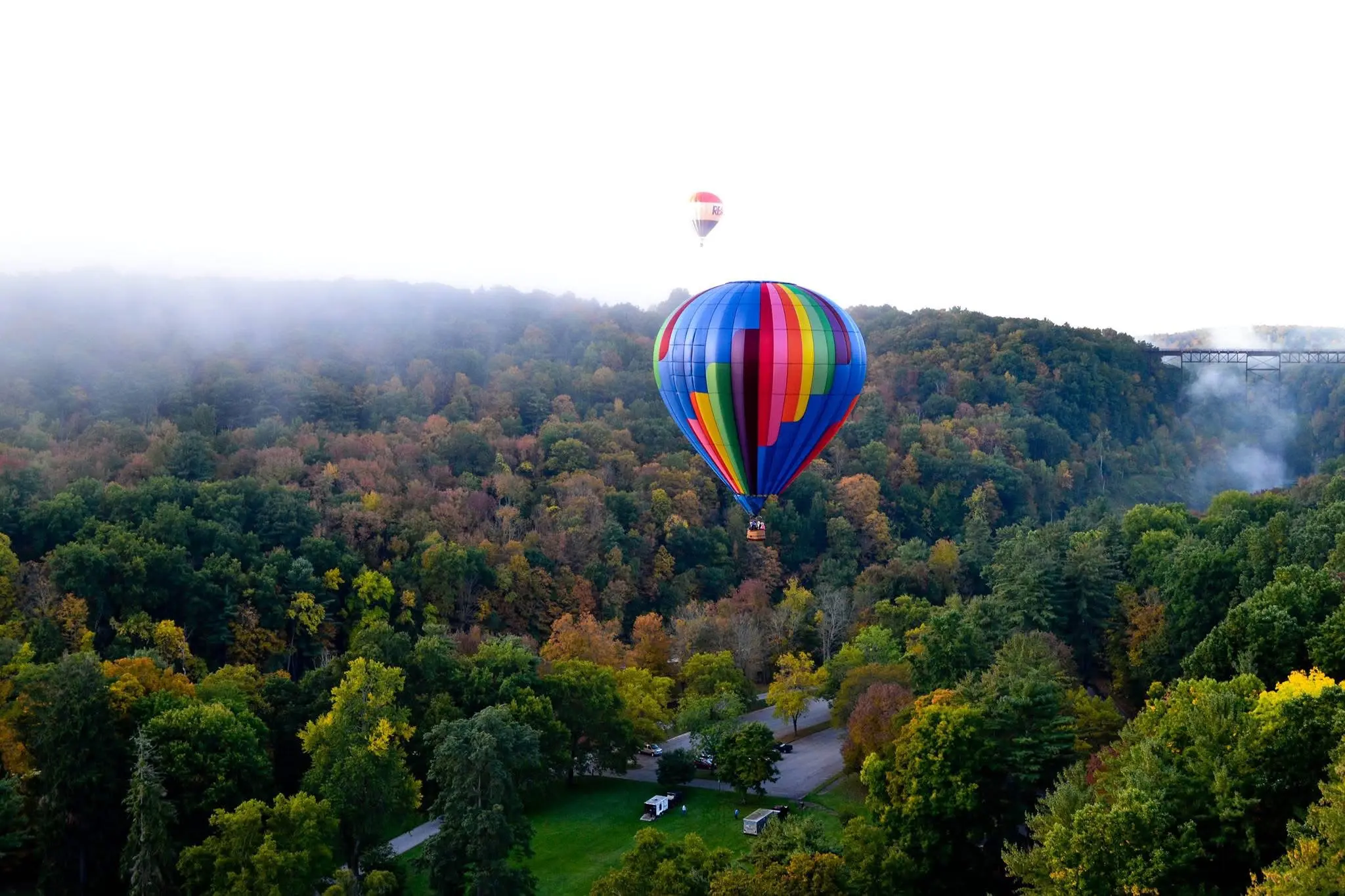 A vibrant hot air balloon floats over a lush forest with autumn colors.