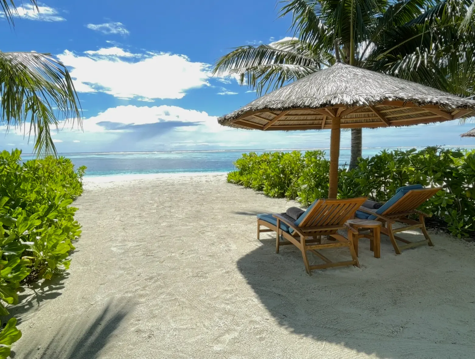 Beach chairs under a thatched umbrella facing the ocean on a sunny day.